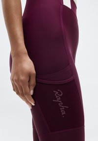 Gros plan sur une main reposant sur la poche latérale d'un legging de sport violet foncé avec le logo "Rapha" sur le tissu de la poche.