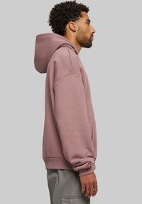 Sudadera de algodón rosa con un bolsillo frontal, mangas largas y capucha ajustable; mostrada de lado sobre un fondo neutro.