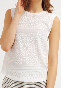 Vit ärmlös topp med blommönster broderier, rund halsringning och texturerad nederkant, kombinerad med randig tyg nedanför.