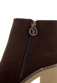 Gros plan d'une botte en daim marron avec fermeture éclair latérale, présentant une languette ronde en métal embossée du logo USPA d'un joueur de polo.