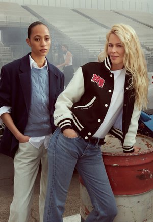 Due donne stanno fianco a fianco. Una indossa una giacca varsity nera e bianca con dettagli rossi; l'altra un maglione blu e un blazer.