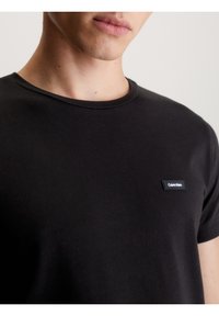 Musta puuvillainen t-paita, jossa pyöreä pääntie ja lyhyet hihat, ja pienellä mustalla Calvin Kleinin logopatchilla rinnassa.