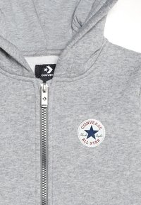 Grijze rits-hoodie gemaakt van een zacht materiaal, met een trekkoordcapuchon en een ronde patch met een blauwe ster en de tekst "Converse All Star".