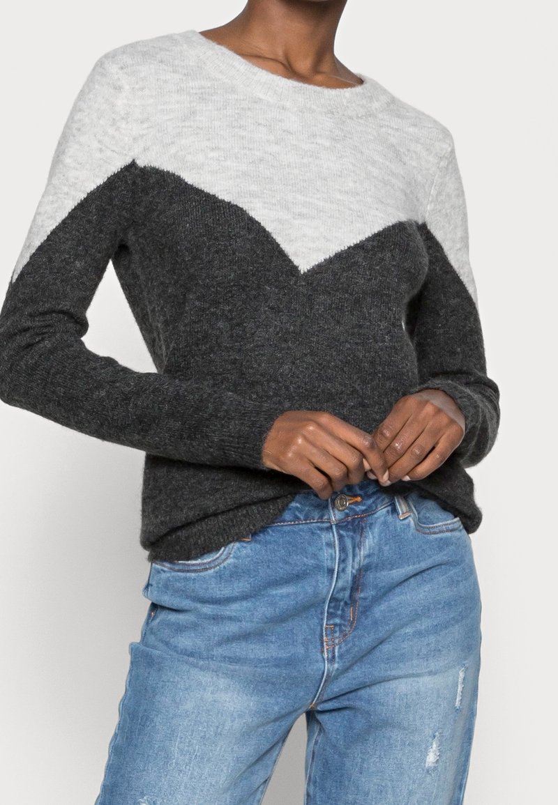 Personne portant un pull en maille à manches longues bicolore avec un motif chevron gris et charbon foncé, associé à un jean bleu clair déchiré.