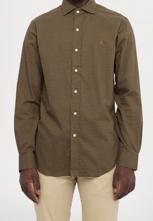 Hombre vestido con una camisa marrón de cuadros con botones y mangas largas, y pantalones beige, de pie frente a un fondo liso.