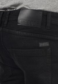 Jeans de mezclilla negros con un parche de cuero en la cinturilla con "INDICODE" en relieve, una etiqueta pequeña adicional en el bolsillo trasero y tela texturizada.