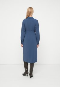 Hosszú ujjú, övvel ellátott ruha sötétkék egyszínű anyagból, gallérral és egyenes szabású alsó résszel, térd fölé érő barna csizmával párosítva.