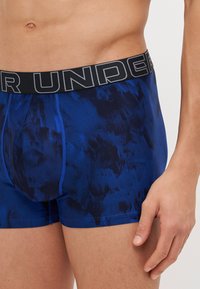 Plave gaćice s uzorkom i crnim elastikom u struku na kojem piše "UNDER" u sivoj boji. Materijal je gladak s suptilnom teksturom.