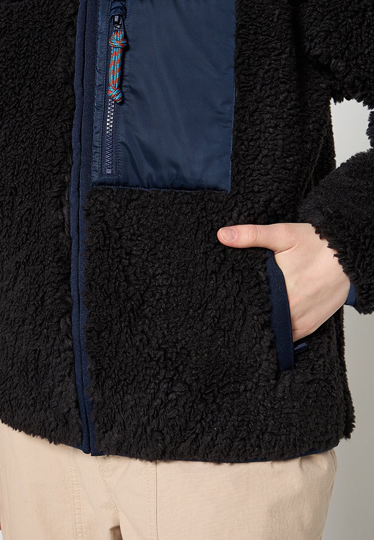 Schwarze Fleecejacke mit strukturierter Oberfläche, ausgestattet mit einem marineblauen Panel und Reißverschluss sowie einer Seitentasche, in die eine Hand gesteckt ist.
