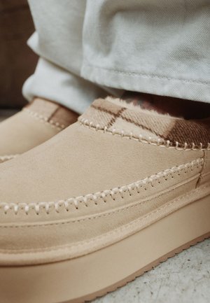 Chaussons mocassins en daim beige avec coutures apparentes et doublure en tissu à carreaux doux, portés avec un pantalon clair.