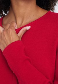 Röd tröja med en mjuk textur, långa ärmar och en vid halsringning, med en synlig hand som försiktigt rör vid halsringningen.