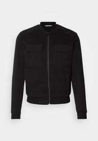 Veste noire avec une fermeture éclair à l'avant, col arrondi et deux poches à rabat sur le devant. Conçue en tissu lisse avec des poignets et un ourlet côtelés.