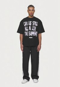 Mladý muž s copánky, oblečený v černém oversized tričku s bílým nápisem "Můžeme se ještě zamilovat toto léto?" a černých kalhotách s bílými pruhy.