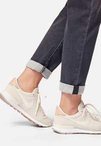 Zapatillas de color beige claro con parte superior de ante, suela blanca y suaves acentos en color marrón claro. Jeans oscuros ajustados con puños en gris claro.