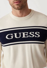 Pulover crem cu un panou bleumarin pe care este inscripționat "GUESS" în alb. Guler rotund cu nervuri și țesătură texturată pe toată suprafața.