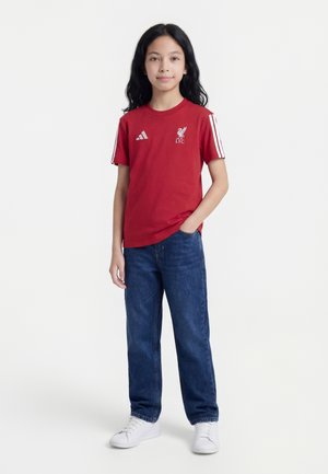 Mladý človek stojaci s rukou v vrecku, oblečený v červenom tričku Liverpool FC, modré džínsy a biele tenisky na jednoduchom pozadí.