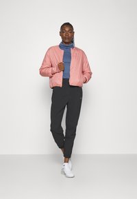 Veste bomber rose clair avec une texture lisse, portée sur un col roulé bleu marine et associée à un pantalon de jogging noir. Baskets blanches présentes.