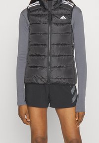 Svart pufferväst med hög krage, dragkedja framtill och horisontell sömnad. Har vita Adidas-stripes på axlarna, som bärs över en grå långärmad tröja.