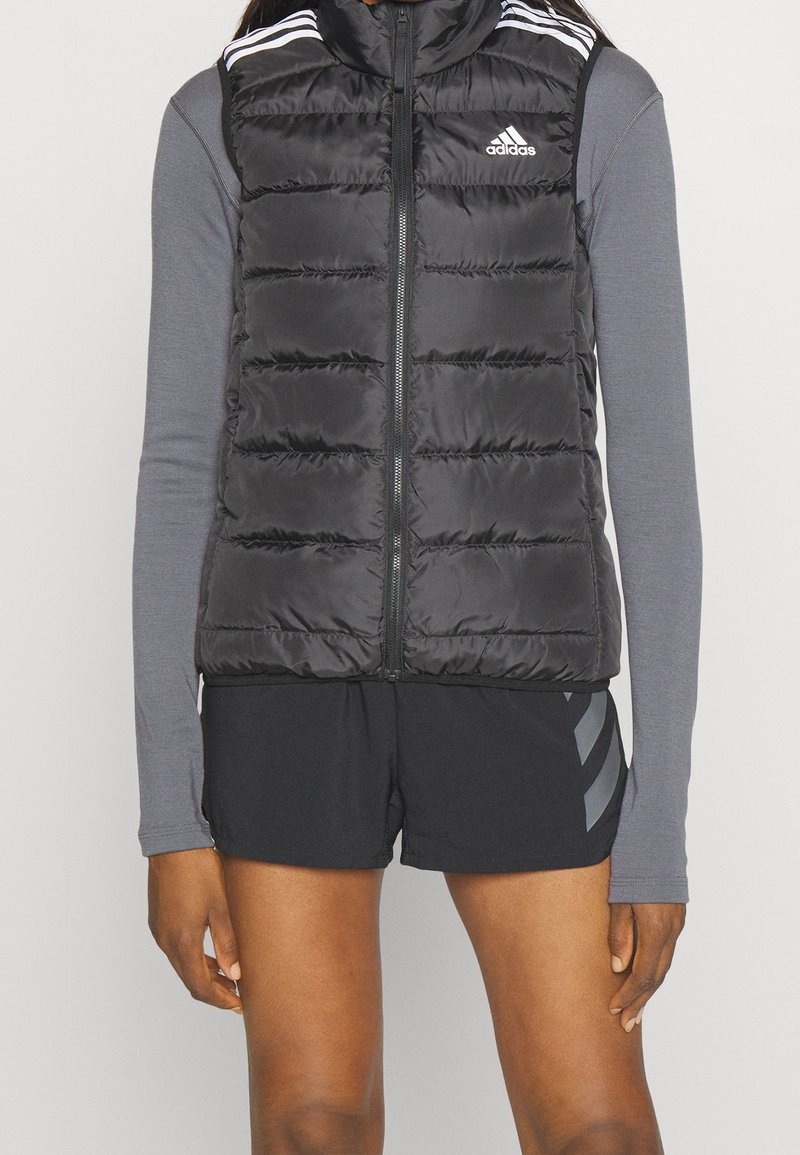 Svart pufferväst med hög krage, dragkedja framtill och horisontell sömnad. Har vita Adidas-stripes på axlarna, som bärs över en grå långärmad tröja.