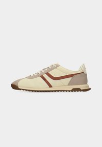 Baskets beige profil bas avec des accents en daim marron, des bandes brunes sur les côtés, des lacets crème et une semelle marron rainurée.
