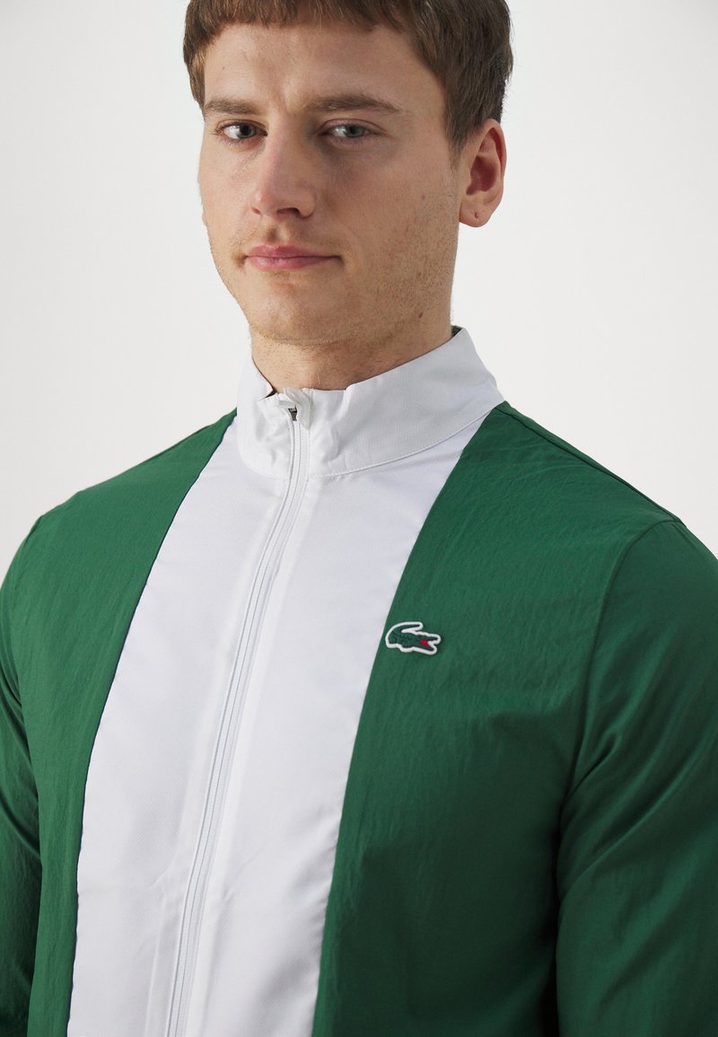 Zielona i biała kurtka zapinana na zamek błyskawiczny z stójką, wykonana z fakturowanej tkaniny, z małym haftowanym logiem na piersi.