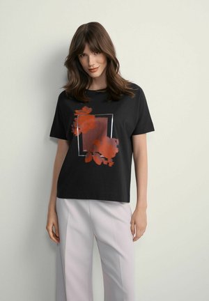 Kobieta nosząca czarną koszulkę z pomarańczowym kwiatowym motywem i jasnoszare spodnie, stoi na tle gładkiej jasnej ściany.