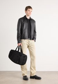 Chaqueta de cuero negra, camiseta negra, vaqueros beige, bolsa negra con asas, zapatos negros, textura suave, diseño moderno, de pie sobre una superficie neutral.