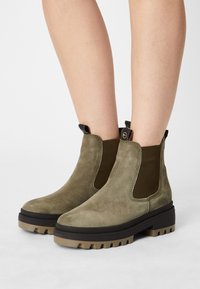 Botas de tobillo de ante verde oliva con paneles laterales elásticos, suela gruesa de relieve y bucles para facilitar su puesta. Textura suave, diseño minimalista.
