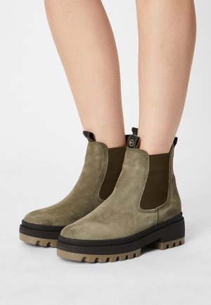 Bottines en suède vert olive avec des panneaux latéraux élastiques, semelle crantée épaisse et languettes pour un enfilage facile. Texture lisse, design minimaliste.