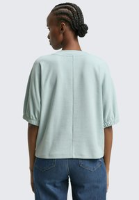 Hellblaue, kurzärmelige Bluse mit rundem Ausschnitt und gerafften Ärmeln. Der Stoff hat eine weiche Textur, und auf der Rückseite befindet sich eine Mittelnaht.