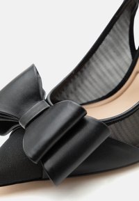 Chaussure plate pour femme noire à bout pointu avec grand nœud en cuir mat et fines panneaux latéraux en mesh sur fond blanc.