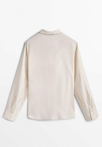 Chemise beige à manches longues avec poignets boutonnés, vue de dos, posée à plat sur une surface blanche.