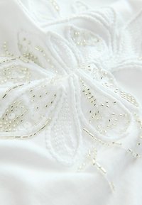Tissu blanc avec une broderie florale complexe en fil blanc et perles argentées, ajoutant texture et détail au design.