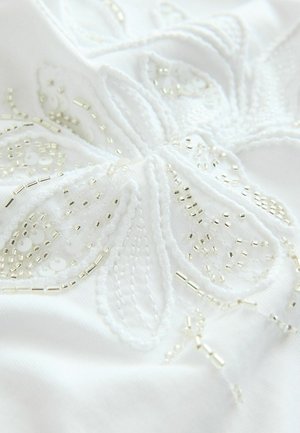 Tissu blanc avec une broderie florale complexe en fil blanc et perles argentées, ajoutant texture et détail au design.