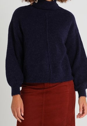 Žena v tmavě modrém roláku s balónovými rukávy a v sukni s vysokým pasem barvy rezavé.