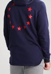 Mörkblå huvtröja med en cirkel av röda stjärnor på ryggen, har en mjuk textur och sidslitsar för en avslappnad passform.