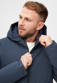 En mand med et velplejet skæg har en marineblå hættejakke på, mens han justerer lynlåsen og kigger tankefuldt til siden.
