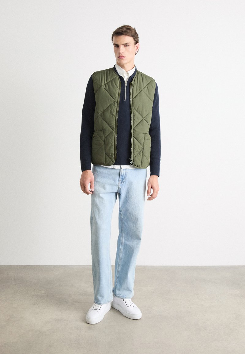 Quilted olivgrüner Weste mit einem Frontreißverschluss, kombiniert mit einem kastanienbraunen Pullover und hellblauen Jeans, getragen mit weißen Turnschuhen.