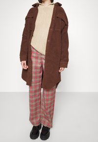 Hnědá fleecová oversized košile, béžový pletený svetr a široké kalhoty s kostkovaným vzorem s červenými akcenty. Černé masivní boty dokončují outfit.