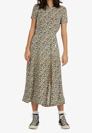 Blumenkleid in Midi-Länge mit kurzen Ärmeln, schwarzer Hintergrund, gelbe und weiße Blumen, seitlicher Schlitz, kombiniert mit schwarzen High-Top-Sneakers und weißen Socken.