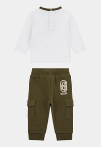 T-shirt : blanche, à manches longues, col olive avec détails de boutons. Pantalons : verts olive, en tissu molletonné, poches cargo, broderie "G" et "GUESS."