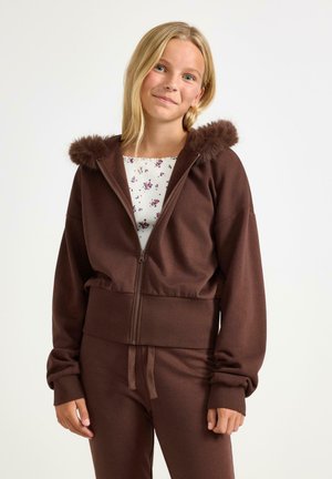 Fille blonde portant un sweat à capuche marron zippé avec bordure en fourrure et pantalon assorti, souriant doucement sur un fond uni.
