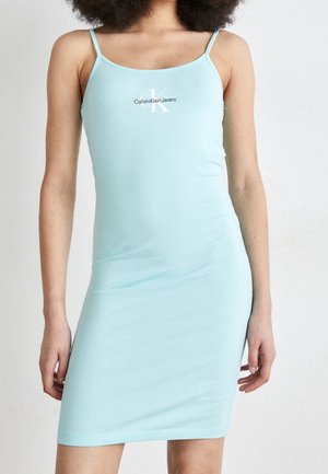 Nainen, joka on pukeutunut vaaleansiniseen, istuvaan Calvin Klein -merkin hihattomaan mekkoon, jossa on ohuet olkaimet ja logo rinnassa, seisoo yksinkertaista taustaa vasten.