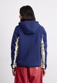 Hættetrøje i marineblå med cremefarvede sidelister; har en glat struktur og kontrastdesign ved håndleddene. Synlig logo på kanten.