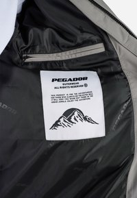 Grå yttertøj med sort foring, har et hvidt mærke med sort tekst og bjerggrafik. Materialet synes glat og vandafvisende.