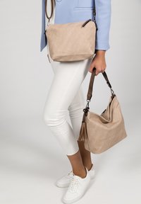 Beige fabric handbag with soft texture, adjustable brown strap, and a tassel accent; paired with a smaller beige crossbody bag.