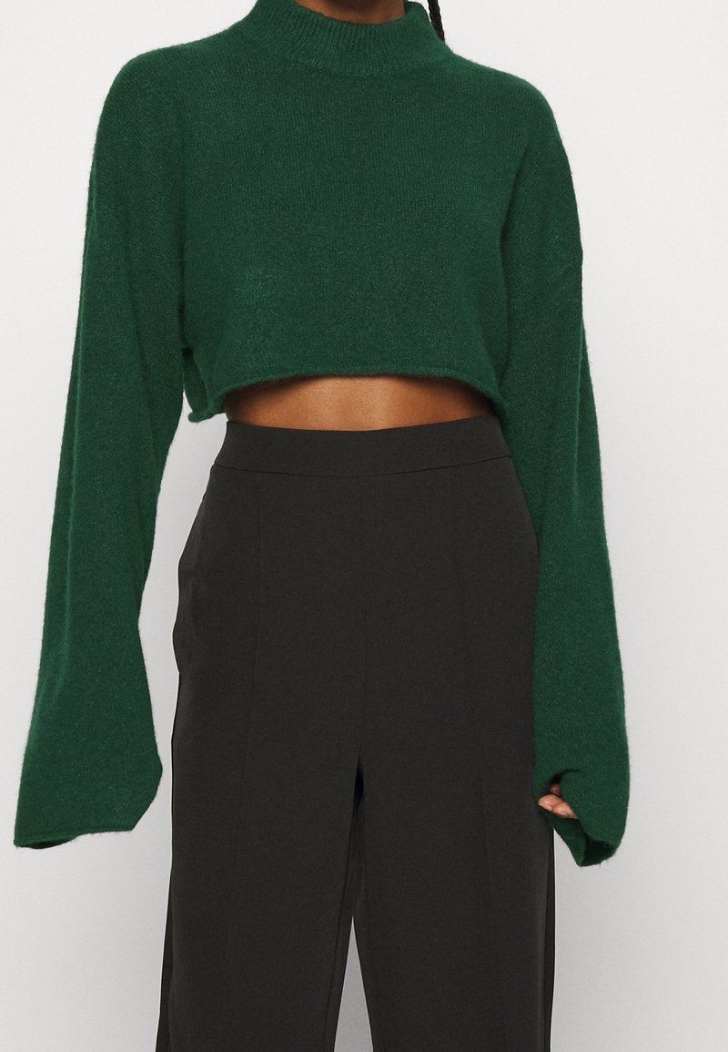 Femme portant un pull court vert foncé à manches longues et un pantalon noir taille haute à jambes larges, sur un fond uni.