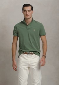 Mlad moški v zeleni polo majici, belih hlačah, rjavem pasu in uri, stoji ob enobarvni svetlo sivi ozadju.