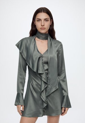 Femme portant une mini-robe en satin gris à manches longues avec des volants et un foulard assorti au cou, debout devant un fond blanc uni.