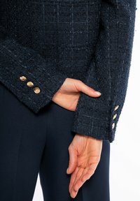 Námořnický tweedový blejzr se strukturovaným povrchem, s mřížkovým vzorem a zlatými knoflíky na rukávech. Tkanina má jemnou, vyvýšenou texturu.